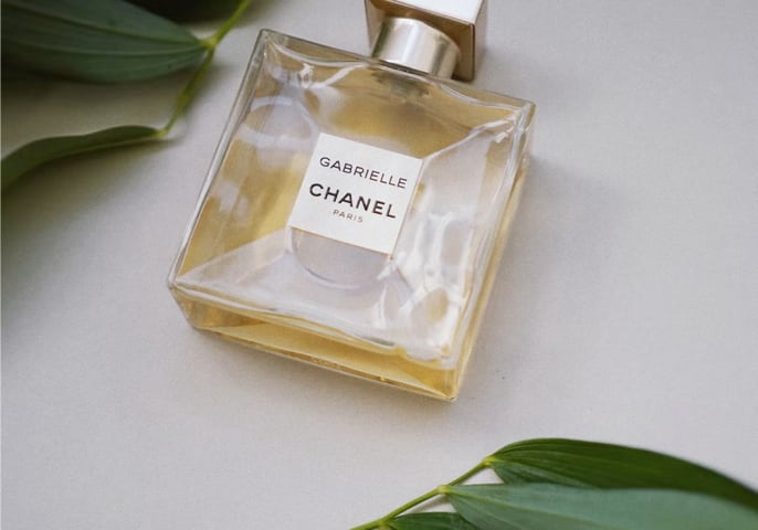 A bottle of Gabrielle Essence Eau De Parfum on a white table, surrounded by leaves.